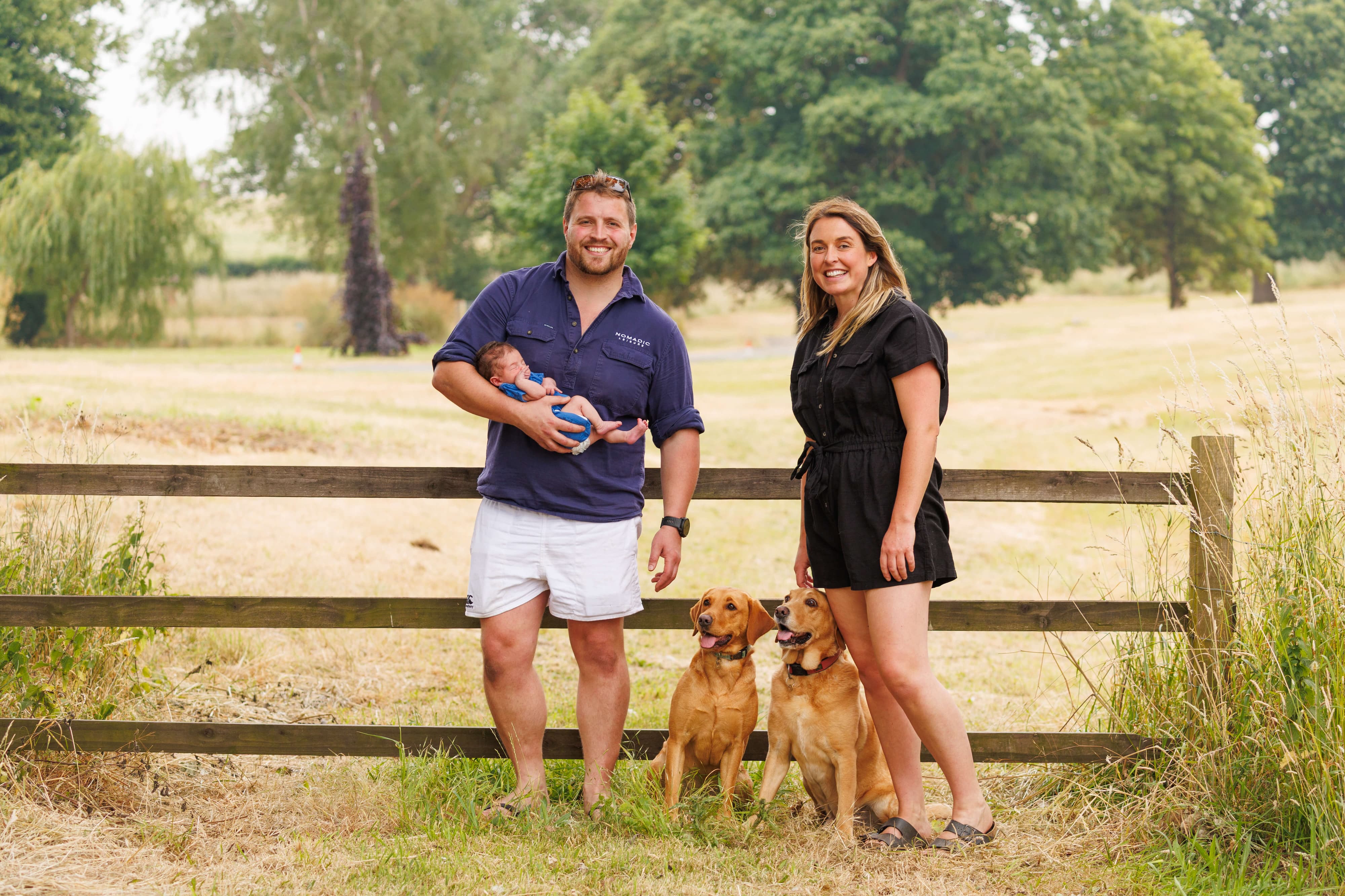 Man and woman with two dogs standing in a field with a wooden fence.