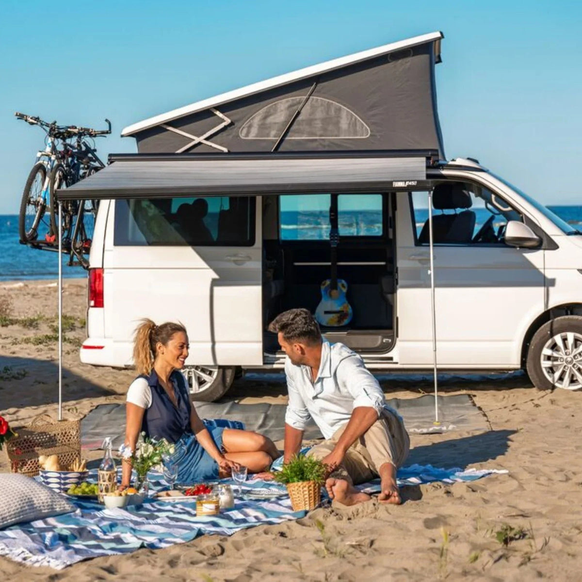 Couple enjoying beach picnic beside campervan with awning and rooftop tent