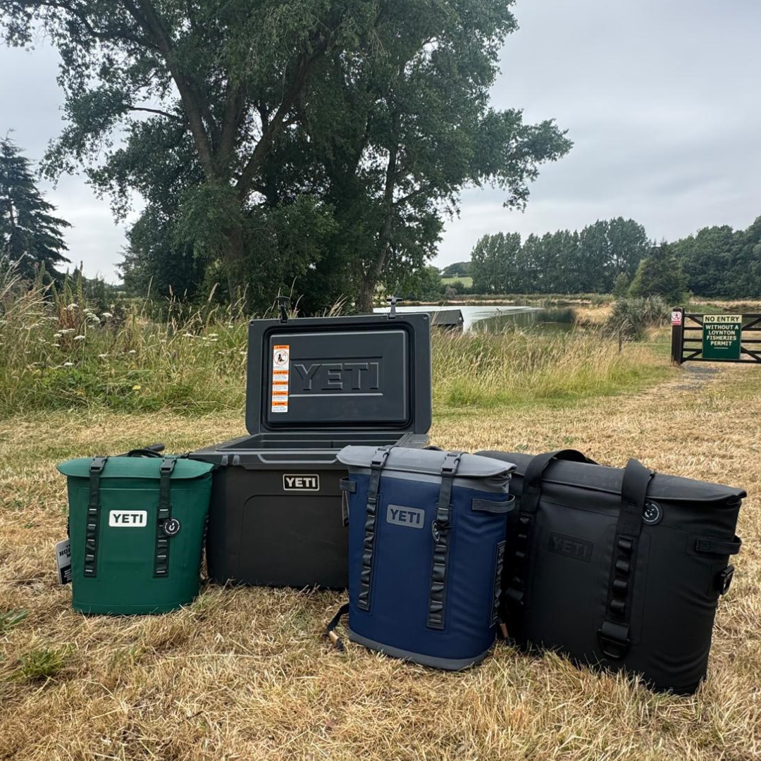 Yeti coolers and soft-sided coolers in green, blue, black on grass near lake for camping, off-grid, marine adventures