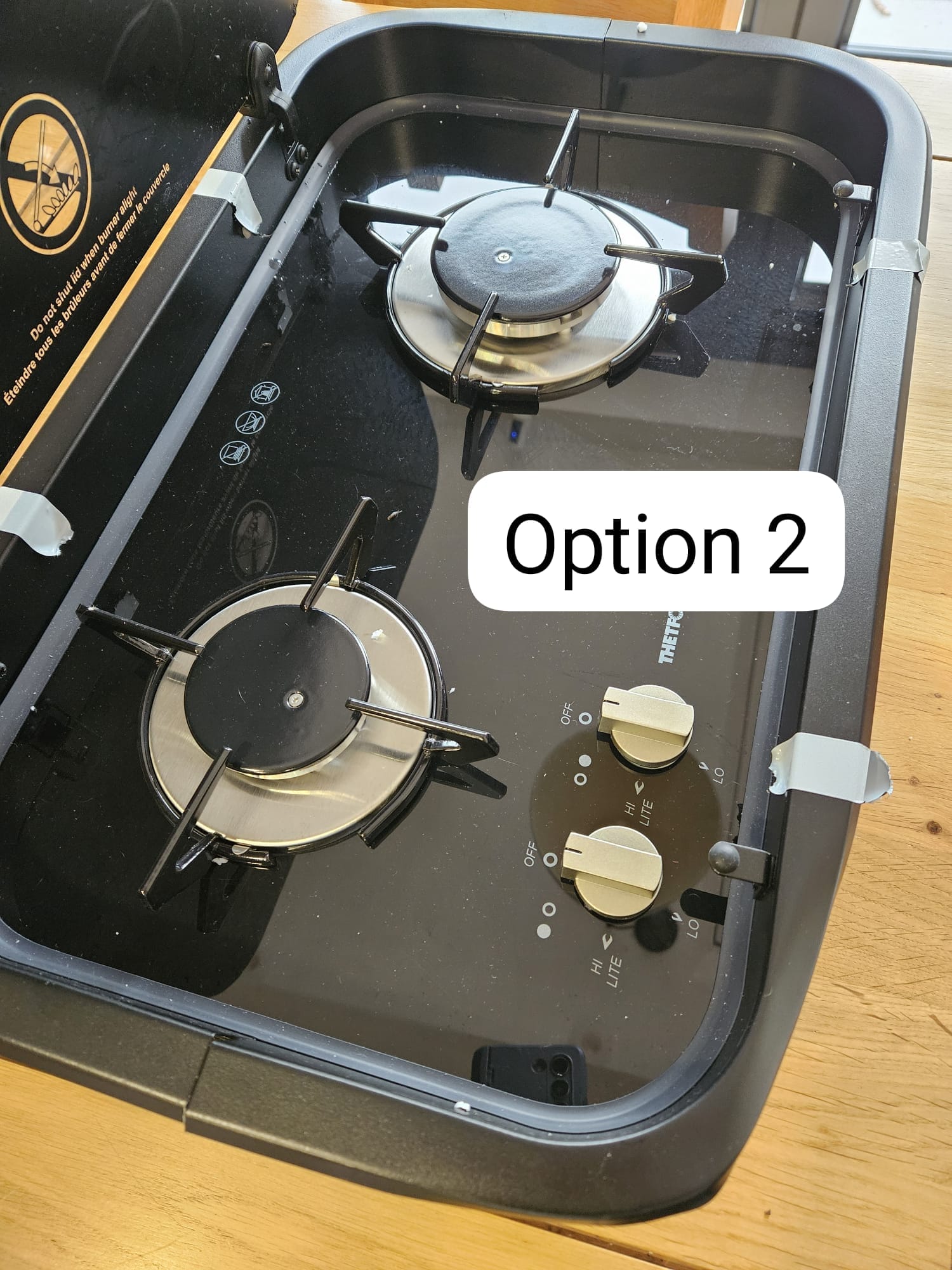 Two-burner campervan gas hob with glass lid and control knobs on wooden surface